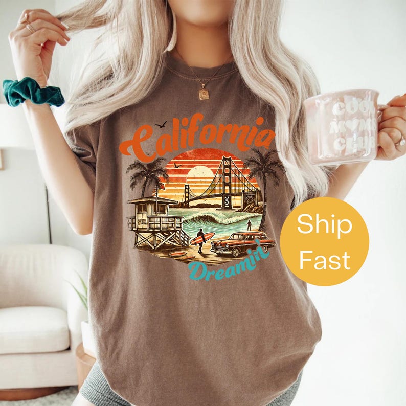 May include: Brown t-shirt with a California graphic featuring the Golden Gate Bridge, a surfer, and a vintage car. The design includes the words "California Dreamin'" in a retro style. The shirt is a relaxed fit.