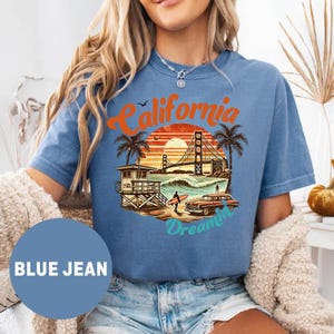 May include: Blue jean t-shirt with a graphic of a California sunset scene. The design includes the Golden Gate Bridge, palm trees, a lifeguard stand, a vintage car, and the words "California Dreamin'" in orange and blue.