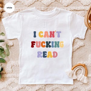 May include: White t-shirt with the text "I CAN'T FUCKING READ" in colourful, retro-style lettering. The shirt is made of a soft material and is suitable for children. The design features a playful and bold statement.
