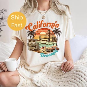 May include: Cream-colored t-shirt featuring a retro California design. The graphic includes the text "California Dreamin'" with a sunset, palm trees, a lifeguard tower, a classic car, and the Golden Gate Bridge.
