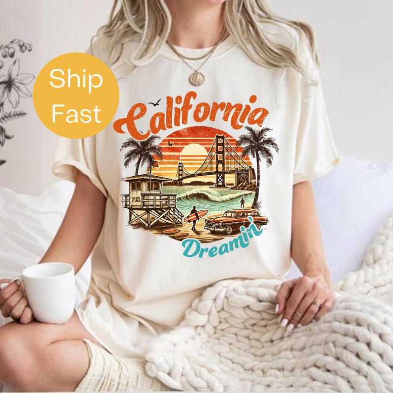 May include: Cream-colored t-shirt featuring a retro California design. The graphic includes the text "California Dreamin'" with a sunset, palm trees, a lifeguard tower, a classic car, and the Golden Gate Bridge.