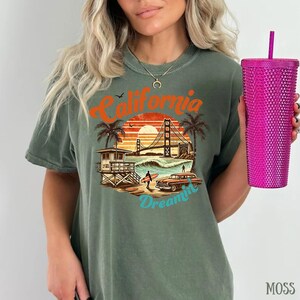 May include: Moss green t-shirt with a California-themed graphic. The design features the Golden Gate Bridge, palm trees, a vintage car, and the words "California Dreamin'". A pink textured tumbler with a straw is held in the hand.