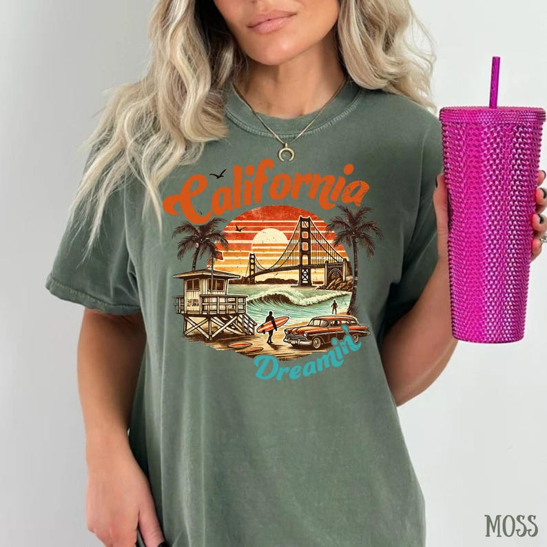 May include: Moss green t-shirt with a California-themed graphic. The design features the Golden Gate Bridge, palm trees, a vintage car, and the words "California Dreamin'". A pink textured tumbler with a straw is held in the hand.