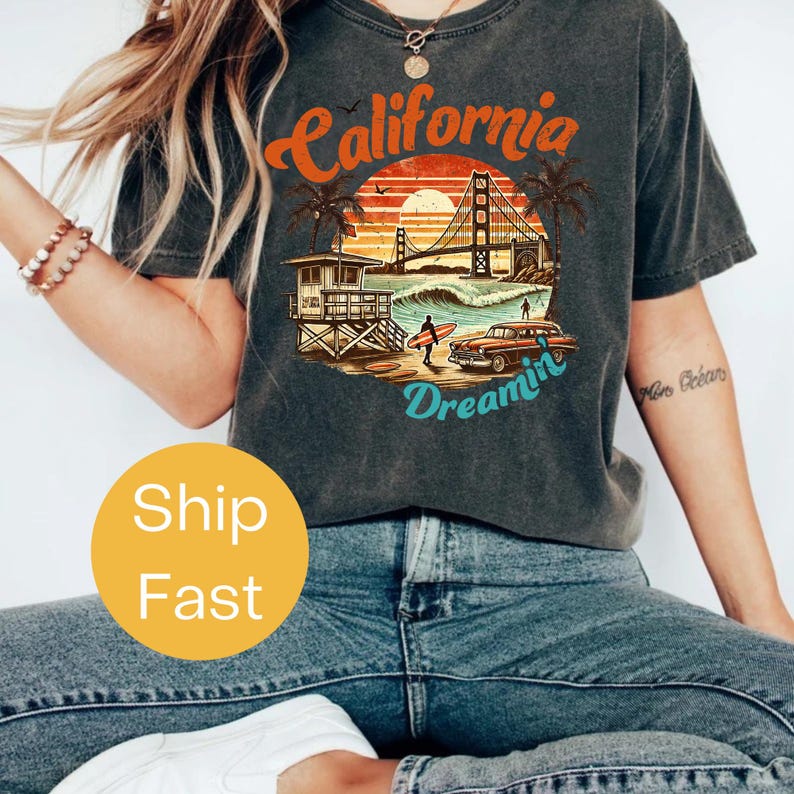 May include: Dark gray t-shirt with a graphic design featuring the words "California Dreamin" in orange and teal. The design includes a sunset, the Golden Gate Bridge, palm trees, a lifeguard stand, a vintage car, and a surfer.