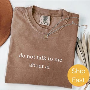 Peut inclure: T-shirt marron Comfort Colors avec le texte "do not talk to me about ai" en blanc. Un collier argenté avec un pendentif est drapé sur le t-shirt. Le t-shirt est plié et possède une étiquette.