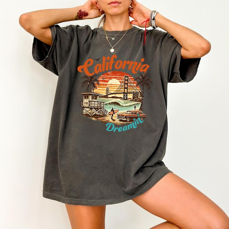 May include: Dark gray t-shirt with a California Dreamin' graphic. The design features a sunset, palm trees, the Golden Gate Bridge, a lifeguard tower, a surfer, and a vintage car. The text "California Dreamin'" is in orange and blue.