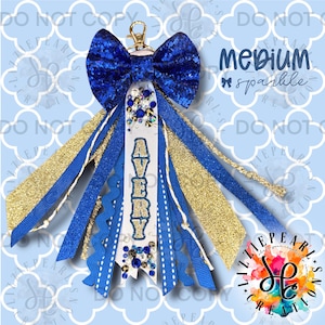 Pode incluir: Um acessório de chaveiro azul e dourado com um laço azul brilhante, fitas e o nome "AVERY" em uma fita branca. O acessório tem um fecho dourado e é decorado com strass. O texto "Medium sparkle" também é visível.