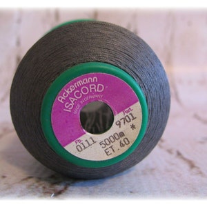May include: A spool of dark gray thread, labeled "Ackermann ISACORD" with a pink and green label. The label includes the text "MADE IN GERMANY" and the numbers "9701" and "0111". The thread is wound around a green spool.