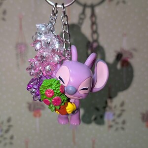 May include: A keychain featuring a lavender-colored cartoon character holding a bouquet of flowers. The character has large ears and closed eyes. The keychain also includes a cluster of pink and clear beads and a silver chain.