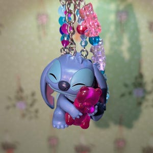 May include: A keychain featuring a blue Stitch figurine holding a pink teddy bear. The keychain has a silver ring and is adorned with colorful beads in shades of pink, blue, and purple. A charming accessory.