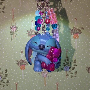 May include: A blue Stitch keychain holding a pink teddy bear. The keychain has a silver ring and is adorned with blue, pink, and clear beads. The background is a green wallpaper with hanging plant illustrations.