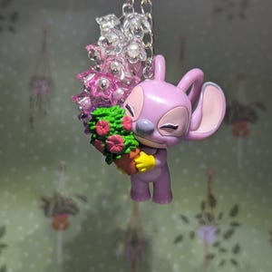 May include: A lavender-colored Angel keychain holding a bouquet of flowers. The keychain features a silver chain and clear and pink beads. The flowers are pink and red, and the pot is brown. The background is a light green wall with a floral pattern.