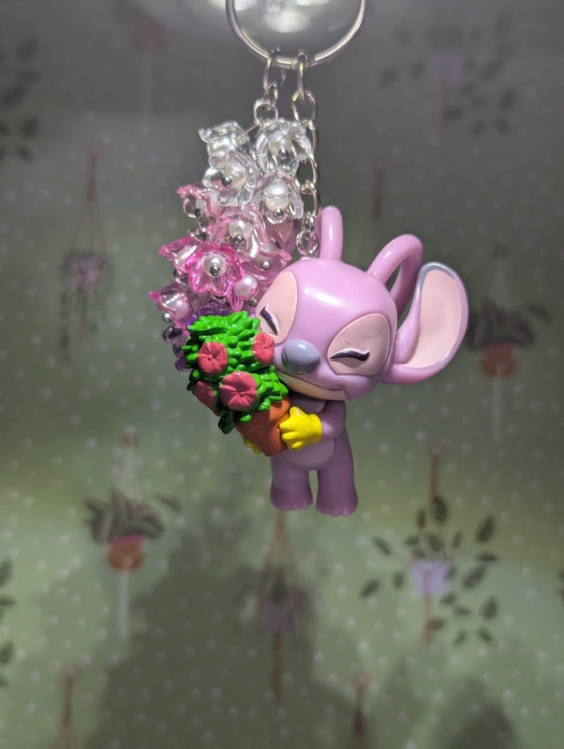 May include: A lavender-colored Angel keychain holding a bouquet of flowers. The keychain features a silver chain and clear and pink beads. The flowers are pink and red, and the pot is brown. The background is a light green wall with a floral pattern.