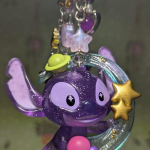May include: A purple glittery Stitch charm with a gold star and a crescent moon. The charm is part of a keychain with other charms, including a green planet, purple stars, and beads.