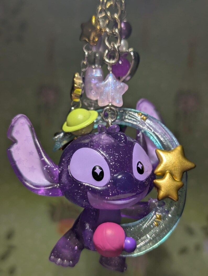 May include: A purple glittery Stitch charm with a gold star and a crescent moon. The charm is part of a keychain with other charms, including a green planet, purple stars, and beads.