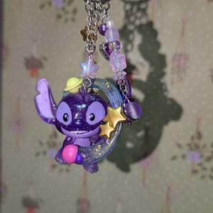 May include: A purple Stitch keychain with a glittery finish, holding a gold star and a crescent moon. The keychain features a beaded chain with star and heart charms, and a silver keyring.