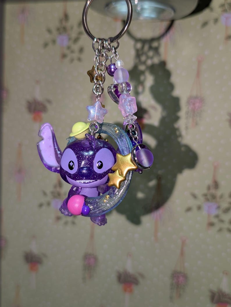 May include: A purple Stitch keychain with a glittery finish, holding a gold star and a crescent moon. The keychain features a beaded chain with star and heart charms, and a silver keyring.
