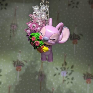 May include: A lavender-colored keychain featuring a cartoon character holding a bouquet of flowers. The character has large ears and is smiling. The bouquet includes pink and red flowers with green foliage. The keychain has a silver-colored chain and a ring.