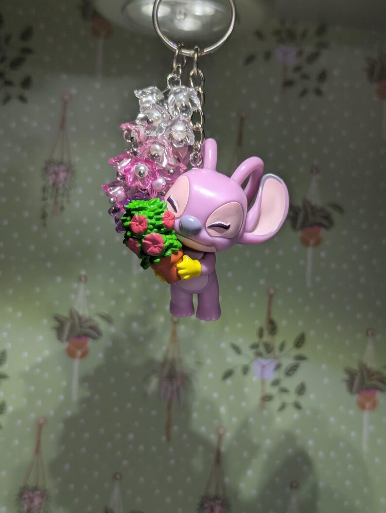 May include: A lavender-colored keychain featuring a cartoon character holding a bouquet of flowers. The character has large ears and is smiling. The bouquet includes pink and red flowers with green foliage. The keychain has a silver-colored chain and a ring.
