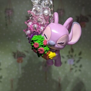 May include: A lavender-colored Angel keychain holding a small potted plant with pink flowers. The keychain features a silver chain and a cluster of clear and pink flower beads. The background is a light green wallpaper with hanging plant designs.