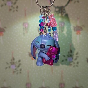 May include: A keychain featuring a blue cartoon character holding a pink bear. The keychain has a silver ring and multiple strands of colorful beads in shades of blue, pink, and clear. The background is a light green wall with a floral pattern.