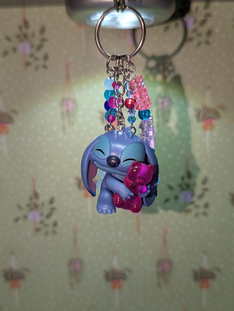 May include: A keychain featuring a blue cartoon character holding a pink bear. The keychain has a silver ring and multiple strands of colorful beads in shades of blue, pink, and clear. The background is a light green wall with a floral pattern.