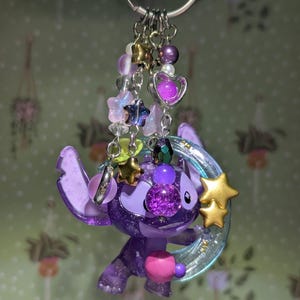 May include: A purple Stitch keychain with a glittery finish, adorned with star and heart charms, and a crescent moon. The keychain features a silver ring and various beaded accents in shades of purple, gold, and clear.