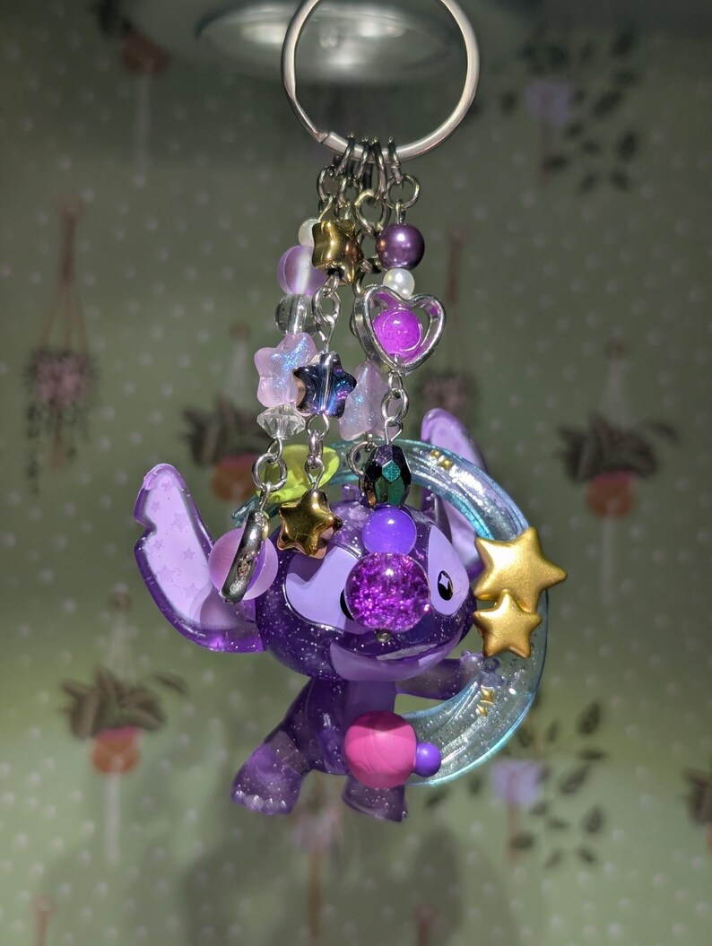 May include: A purple Stitch keychain with a glittery finish, adorned with star and heart charms, and a crescent moon. The keychain features a silver ring and various beaded accents in shades of purple, gold, and clear.