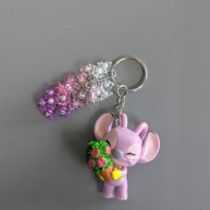 May include: A keychain featuring a lavender-colored character holding a green and pink floral arrangement. The keychain also includes a beaded floral charm in shades of pink, purple, and silver. The background is a solid gray.