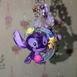 May include: A purple glittery Stitch keychain with a pink ball, a yellow planet, and a gold star. The keychain has a silver ring and is adorned with purple and white beads and star charms.