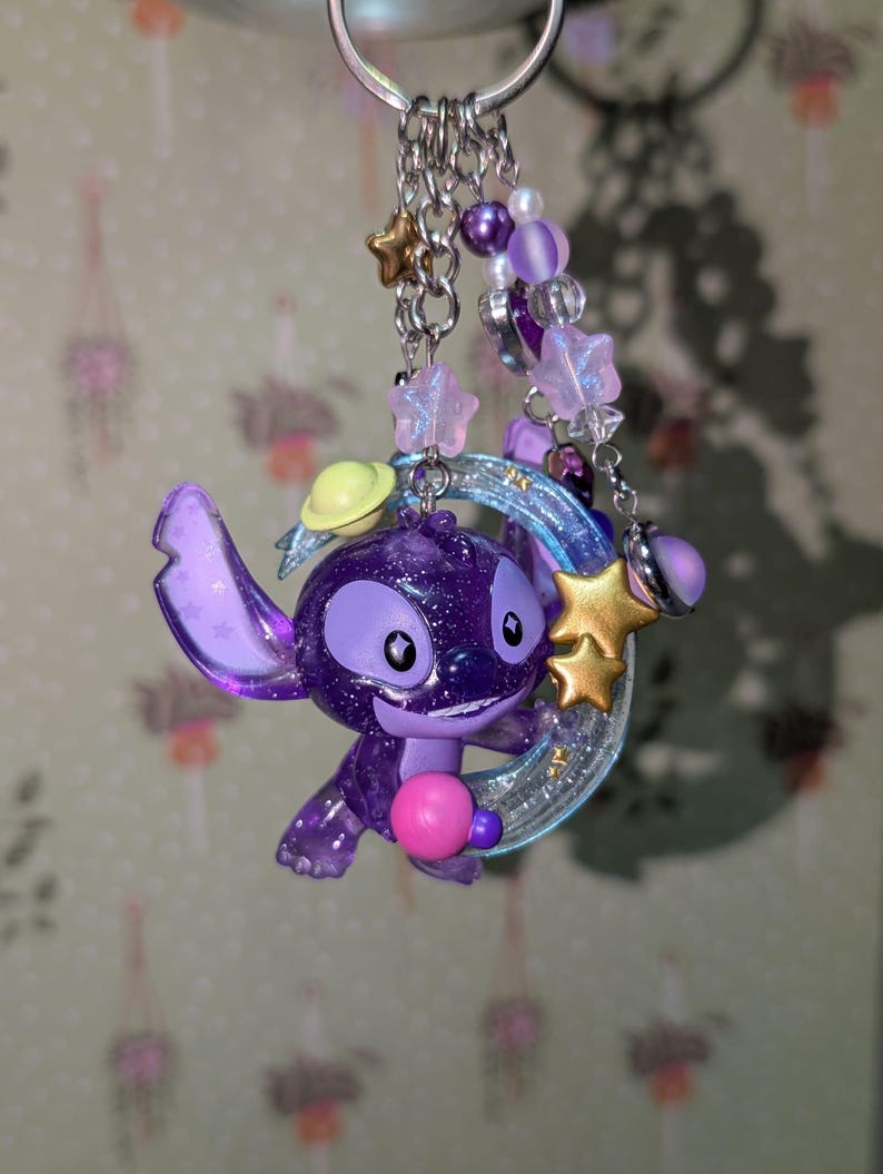 May include: A purple glittery Stitch keychain with a pink ball, a yellow planet, and a gold star. The keychain has a silver ring and is adorned with purple and white beads and star charms.
