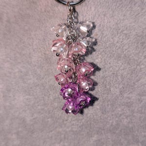 May include: A floral-themed keychain with a silver ring. The charm features a cascade of translucent, flower-shaped beads in shades of pink and purple, accented with small, pearl-like beads. The keychain is suspended from a silver chain.