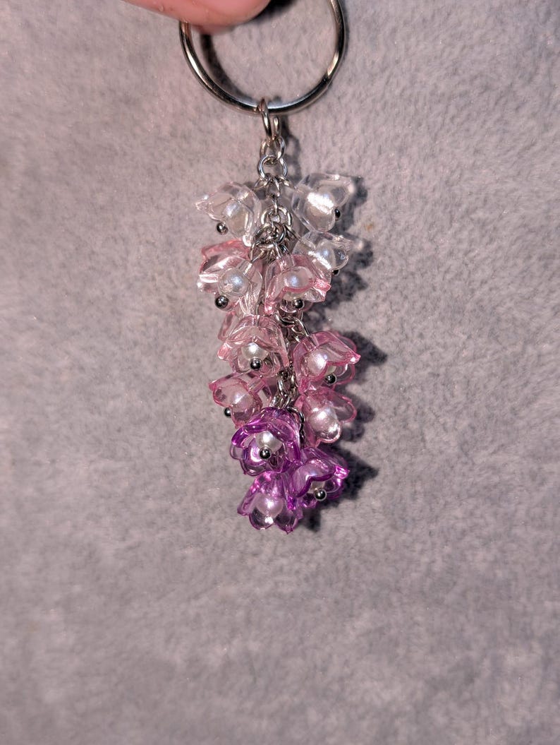 May include: A floral-themed keychain with a silver ring. The charm features a cascade of translucent, flower-shaped beads in shades of pink and purple, accented with small, pearl-like beads. The keychain is suspended from a silver chain.