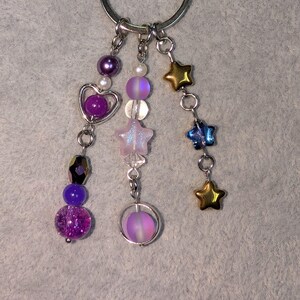 May include: A silver keychain with multiple dangling charms. Charms include purple and iridescent beads, a heart, a star, and gold and blue star-shaped charms. The background is a light gray fabric.