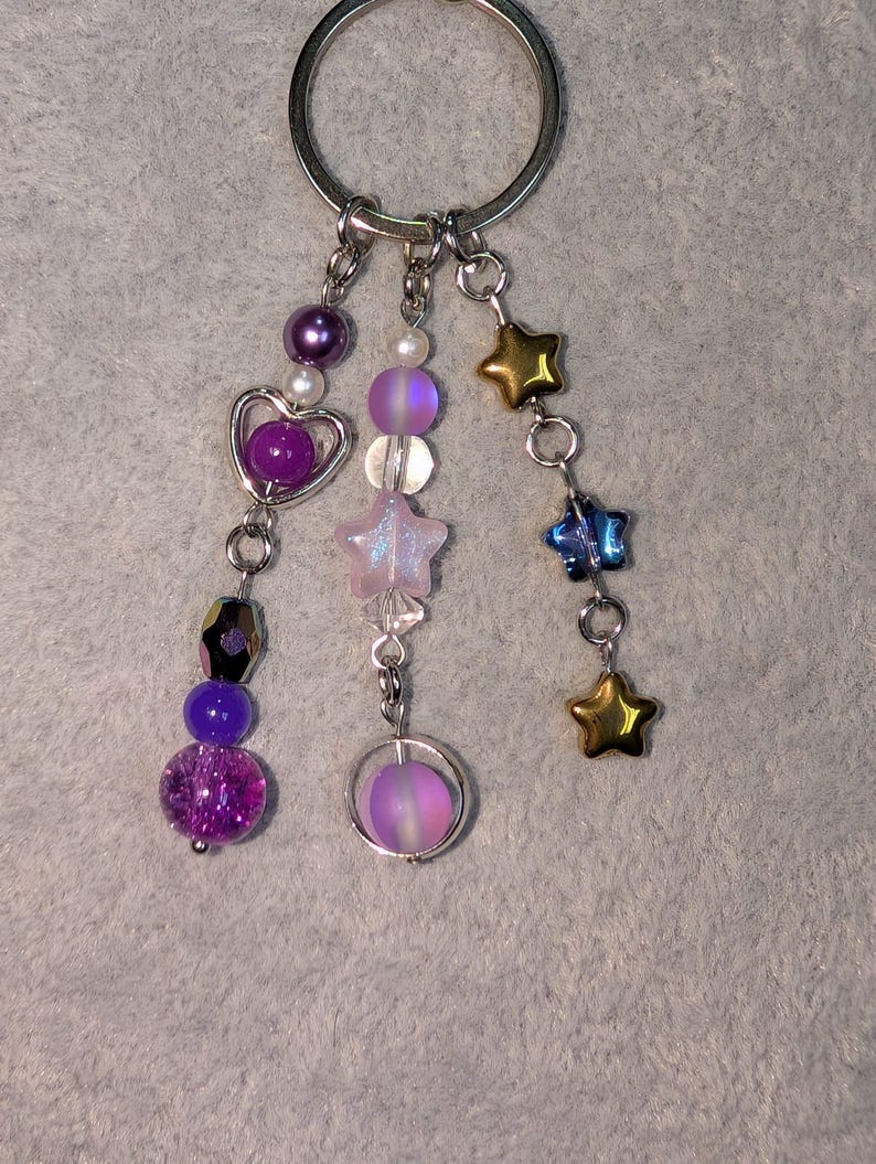 May include: A silver keychain with multiple dangling charms. Charms include purple and iridescent beads, a heart, a star, and gold and blue star-shaped charms. The background is a light gray fabric.