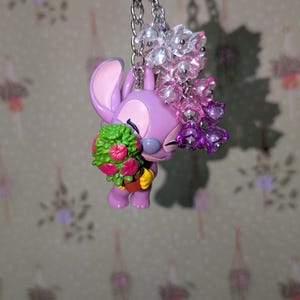 May include: A purple Angel keychain holding a bouquet of flowers. The keychain features a silver chain and a cluster of clear and pink beads. The flowers are green and pink. The background is a floral pattern.