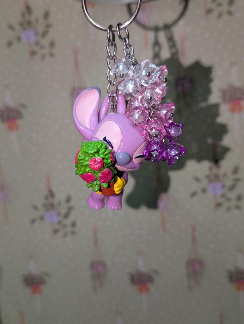 May include: A purple Angel keychain holding a bouquet of flowers. The keychain features a silver chain and a cluster of clear and pink beads. The flowers are green and pink. The background is a floral pattern.