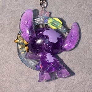 May include: A purple glittery keychain featuring a cartoon character with large ears and a crescent moon design. The character is a shade of purple with glitter accents. The keychain has a silver chain.