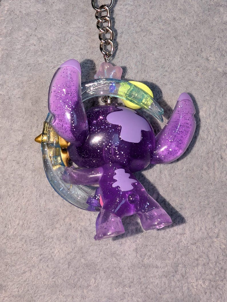 May include: A purple glittery keychain featuring a cartoon character with large ears and a crescent moon design. The character is a shade of purple with glitter accents. The keychain has a silver chain.