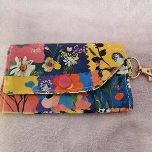 Op de afbeelding: Een kaarthouder met bloemenprint met een roze knoopsluiting en een goudkleurige sluiting. De stof heeft een kleurrijk patroon van bloemen in roze, blauw, geel en wit.