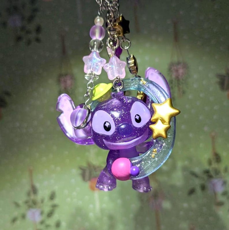 May include: A purple Stitch figurine with glitter, holding a translucent blue crescent moon with gold stars. The charm is attached to a silver chain with pink and purple star-shaped beads and other decorative beads. A pink ball is held in Stitch's hand.