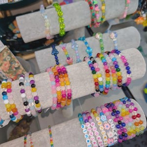 Könnte beinhalten: Eine Präsentation von bunten Perlenarmbändern in verschiedenen Designs. Die Armbänder weisen eine Vielzahl von Perlenformen und -farben auf, darunter Rosa, Lila, Grün und Blau. Einige Armbänder haben Buchstabenperlen, die Wörter buchstabieren.