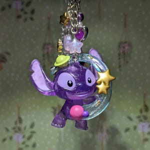 May include: A purple glittery Stitch keychain with a gold star and a crescent moon. The keychain has a silver chain with purple beads, stars, and hearts. The Stitch figure is holding a pink ball.