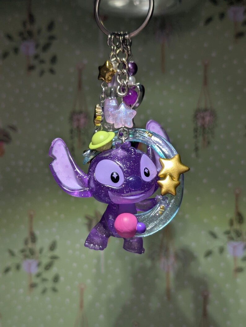 May include: A purple glittery Stitch keychain with a gold star and a crescent moon. The keychain has a silver chain with purple beads, stars, and hearts. The Stitch figure is holding a pink ball.