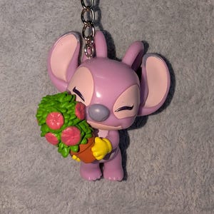 May include: A lavender-colored Angel keychain figurine holding a green and pink floral arrangement in a brown pot. The figurine has large ears, yellow gloves, and closed eyes. A silver chain is attached to the top.