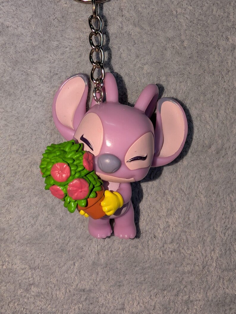 May include: A lavender-colored Angel keychain figurine holding a green and pink floral arrangement in a brown pot. The figurine has large ears, yellow gloves, and closed eyes. A silver chain is attached to the top.