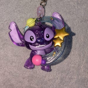 May include: A purple glittery Stitch keychain with a yellow planet hat, holding a pink sphere. The keychain has a silver chain and a light purple bead. Two gold stars and a blue crescent moon are behind Stitch.