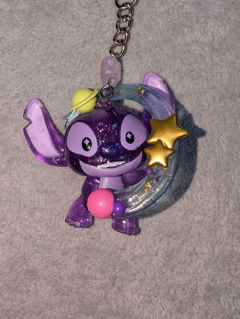 May include: A purple glittery Stitch keychain with a yellow planet hat, holding a pink sphere. The keychain has a silver chain and a light purple bead. Two gold stars and a blue crescent moon are behind Stitch.