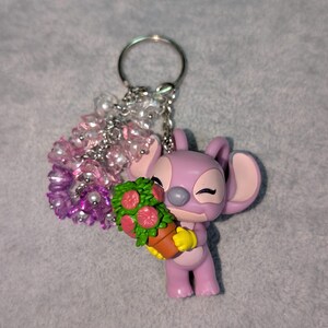 May include: A lavender-colored keychain featuring a cartoon character holding a flower pot with red flowers. The keychain has a silver ring and a cluster of pink and white beads. The character has large ears and closed eyes.