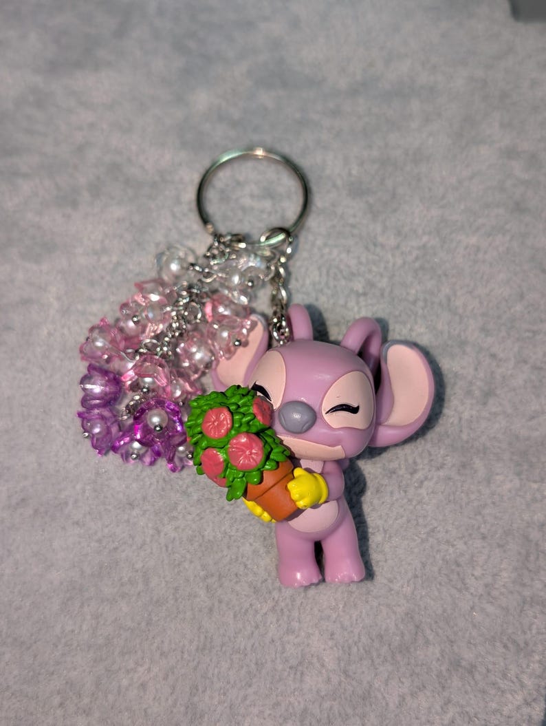 May include: A lavender-colored keychain featuring a cartoon character holding a flower pot with red flowers. The keychain has a silver ring and a cluster of pink and white beads. The character has large ears and closed eyes.